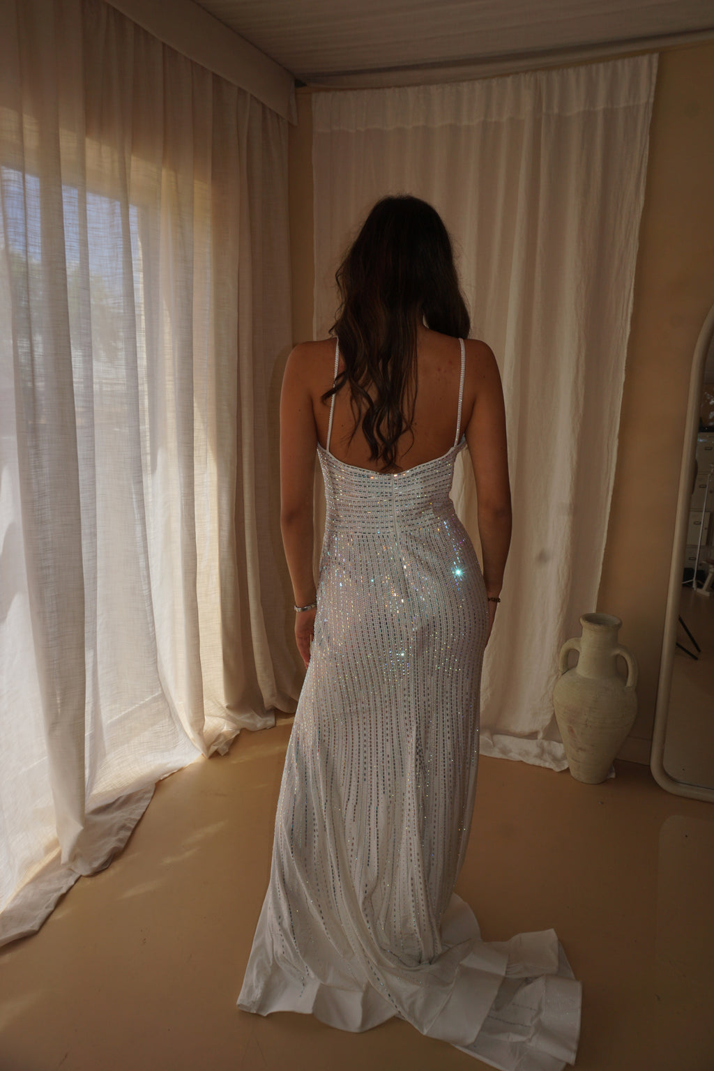 A woman viewed from behind wearing a sleeveless, floor-length white gown covered in shimmering sequins, standing in a softly lit room with cream curtains and a beige floor.