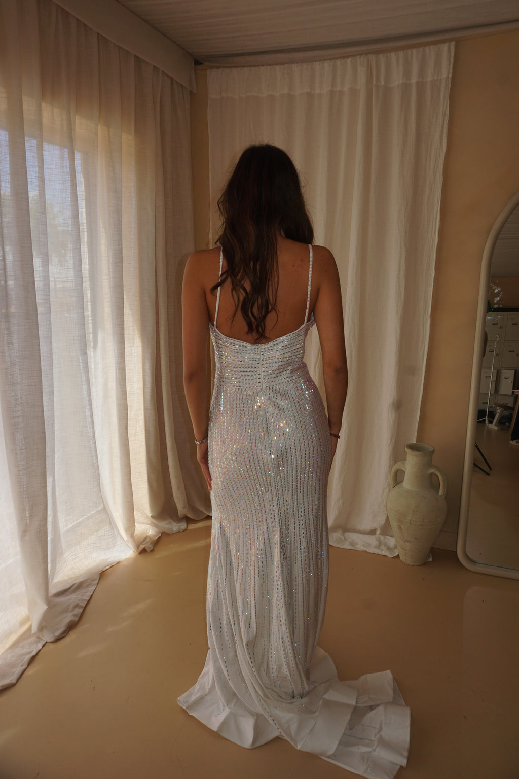 A woman viewed from behind wearing a sleeveless, floor-length white sequined gown with a low open back and a long train, standing in a softly lit room with cream curtains and a mirror.