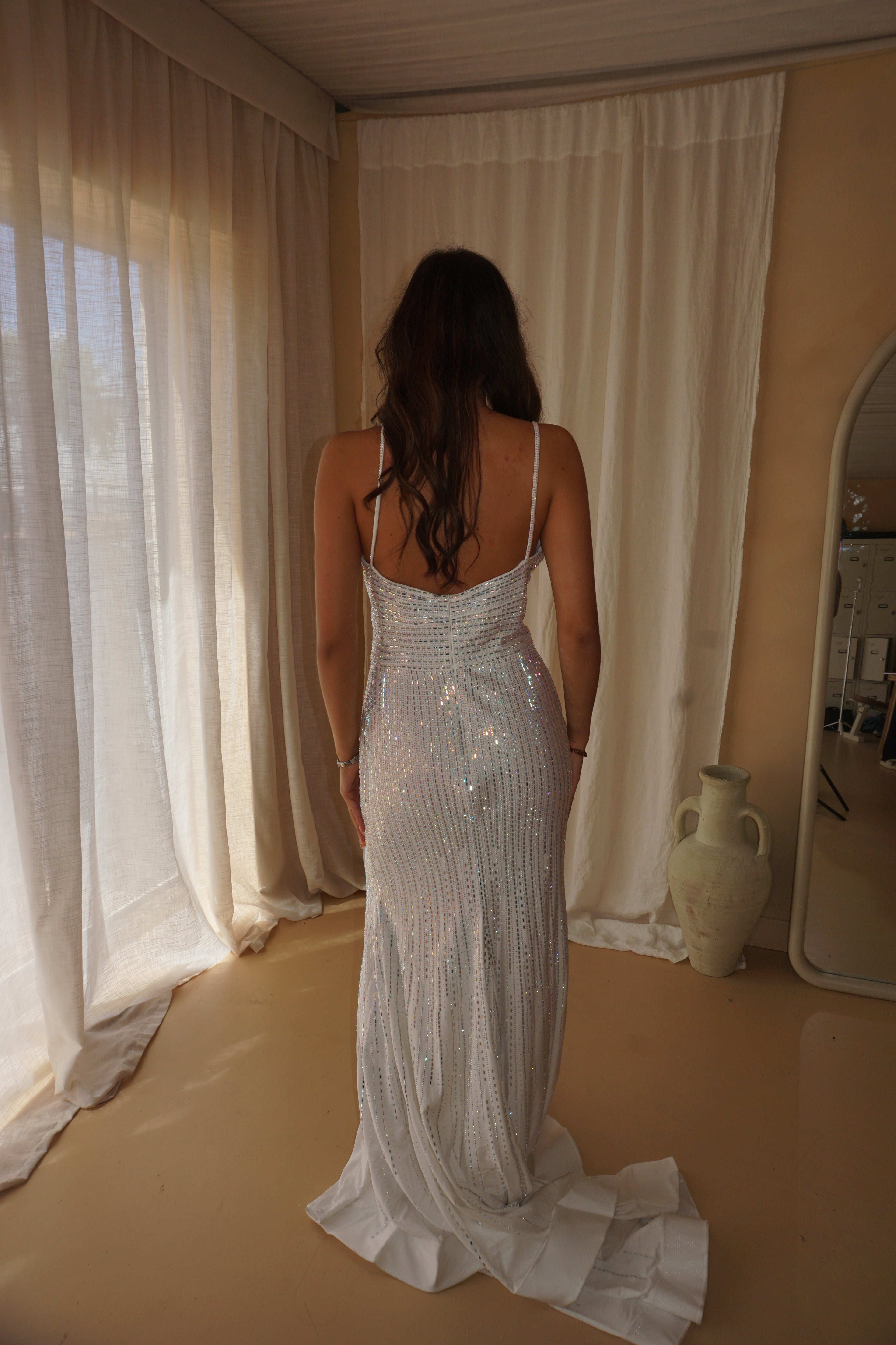 A woman viewed from behind wearing a sleeveless, floor-length white sequined gown with a low open back and a long train, standing in a softly lit room with cream curtains and a mirror.