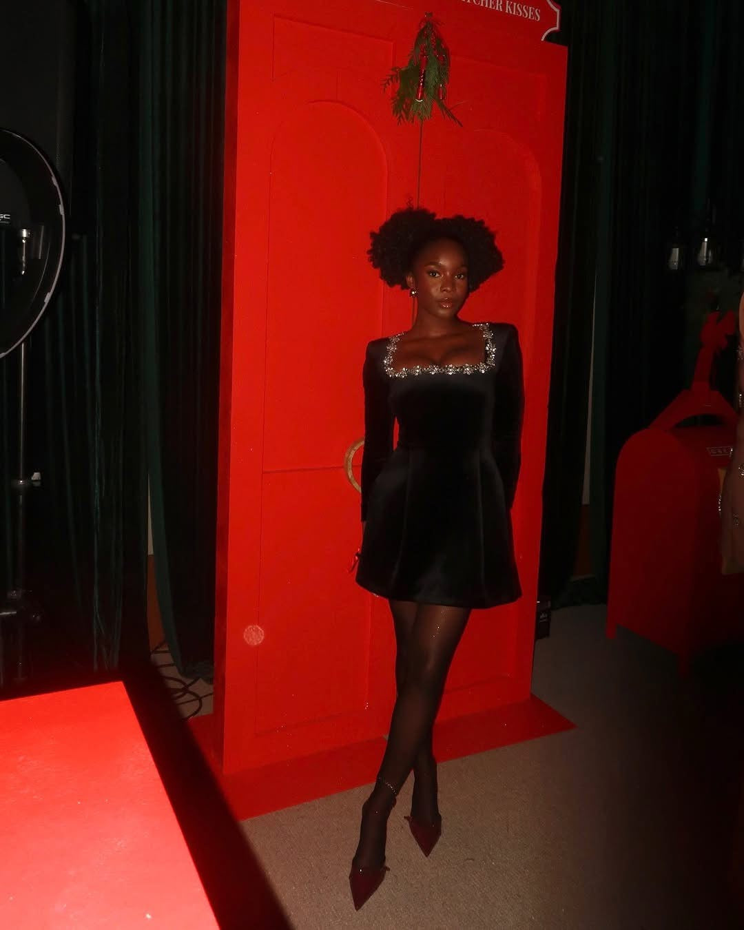 Woman in a black dress standing against a red wall with a fan and coat rack in the background.