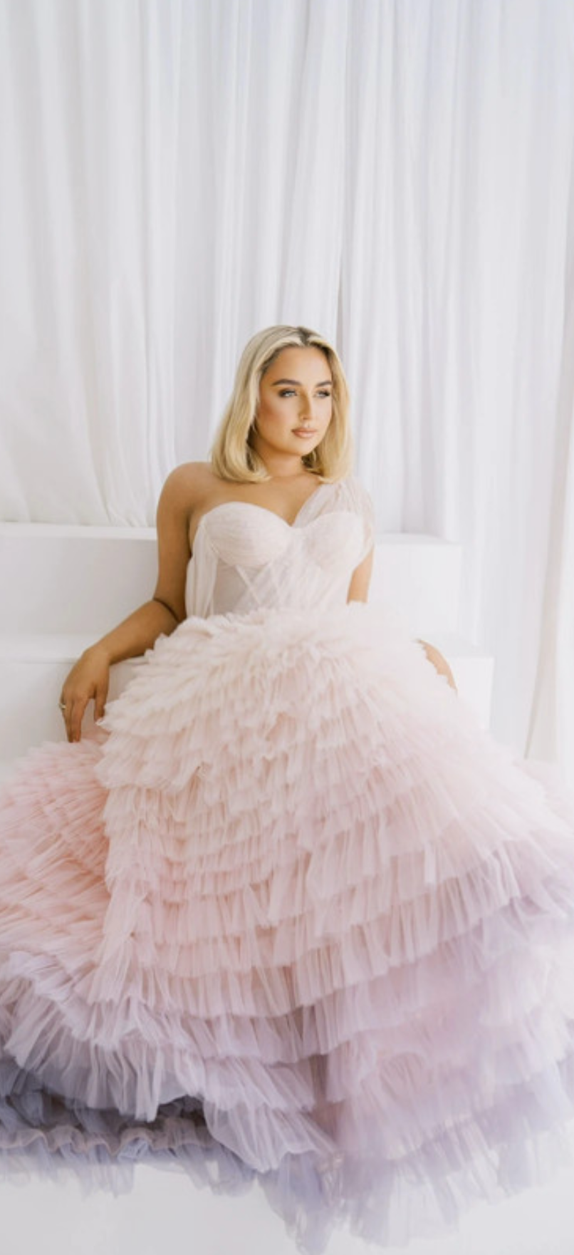 Woman in a pink ruffled gown sitting on a white couch against a white curtain.