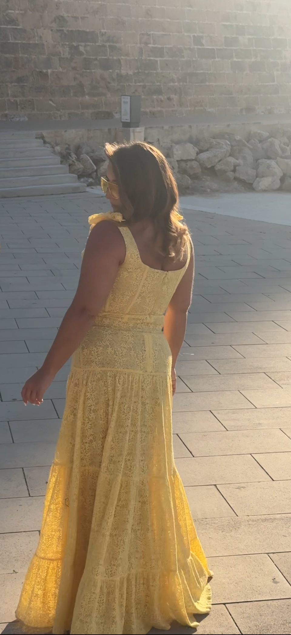 Woman in a yellow dress standing on a paved area with a stone wall in the background.