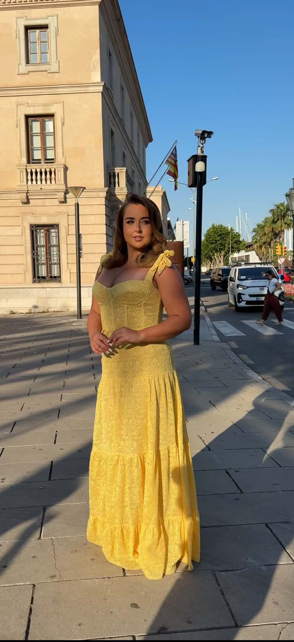 Woman in a yellow dress standing on a street with buildings and cars in the background.